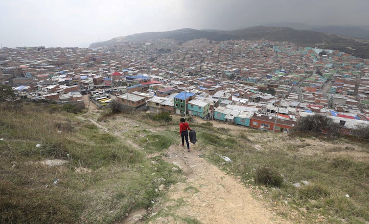 A woman walks along a path towards a crowded urban area seen at the foot of the hills she's walking on.
