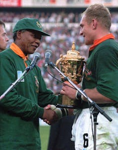 A Black man wears a green ball cap while shaking the hands of a white man holding a trophy.