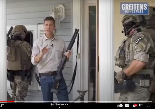 A white man wearing blue jeans is is carrying a high-powered rifle as he stands near three commandos dressed in military gear and ready to enter a house.