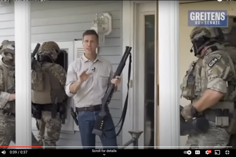 A white man wearing blue jeans is is carrying a high-powered rifle as he stands near three commandos dressed in military gear and ready to enter a house.