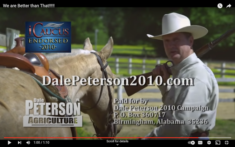 A white man wearing a white cowboys has a rifle on his shoulder as he stands near a horse.