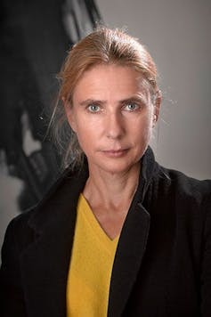 woman with hair pulled back, wearing black jacket and yellow t-shirt, looks seriously at camera