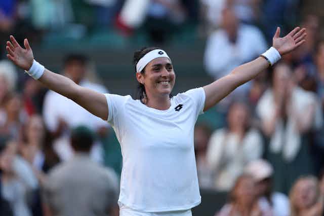 A woman in tennis clothes and sweatbands on her head and wrists smiles as she stretches both arms out wide.