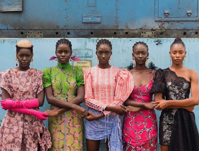 Five Nigerian women models stand against a blue wall in brightly coloured clothing.