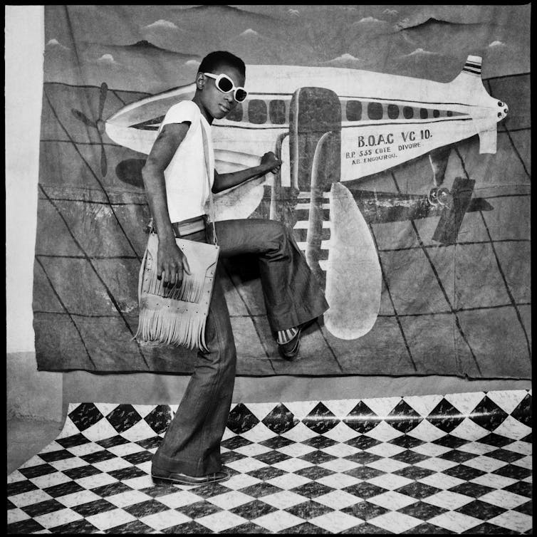 Black and white image of a model in sunglasses pretending to be on a cartoon plane.