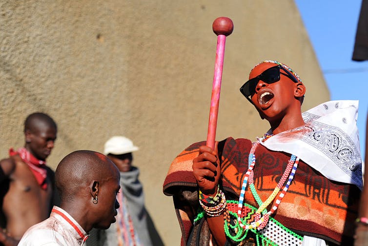 A young man in sunglasses sings and holds a baton.