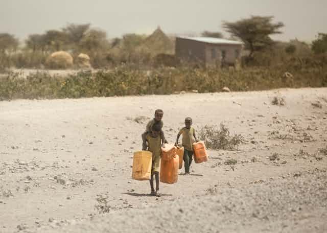 three children walk through dusty ground carrying water containers 