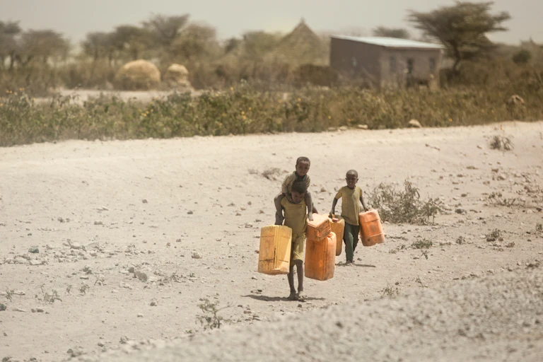 three children walk through dusty ground carrying water containers