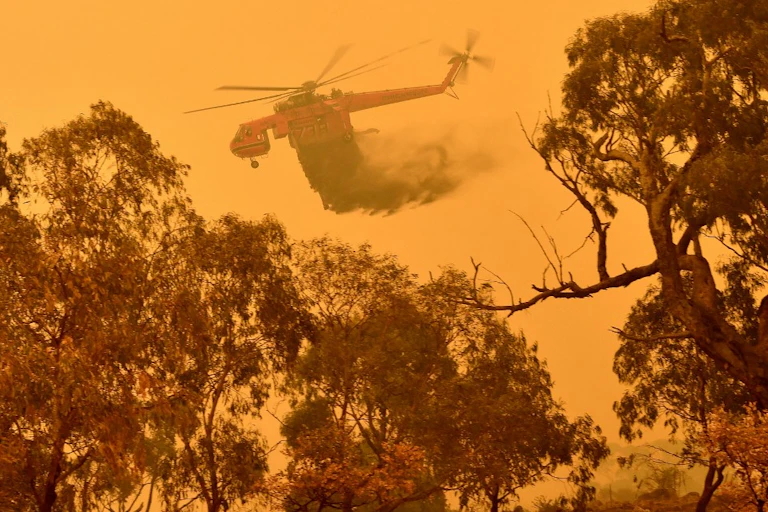 Un hélicoptère bombardier d'eau tente d'éteindre le feu de brousse dans un ciel orange.