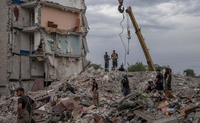Rescue workers stand on the rubble of a destroyed apartment building. A couch is seen sitting a few storeys above them. 