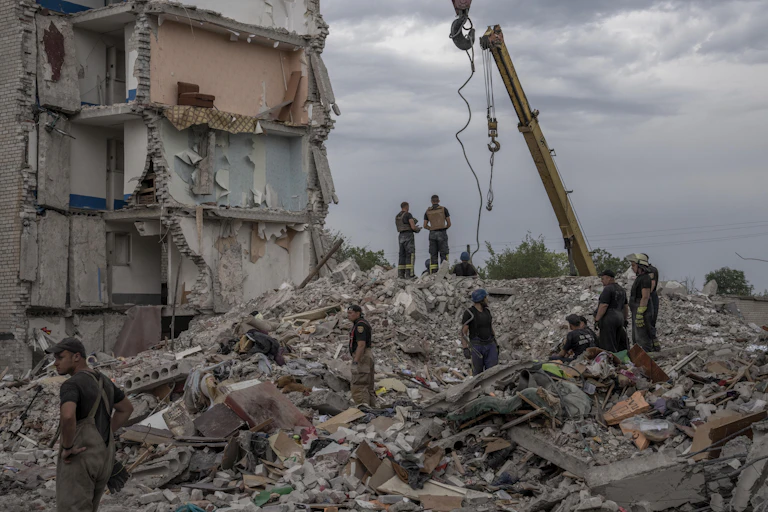 Rescue workers stand on the rubble of a destroyed apartment building. A couch is seen sitting a few storeys above them.