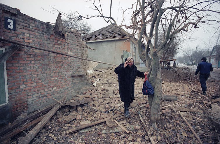 An elderly woman weeps as she stands near rubble with her hand on a tree.