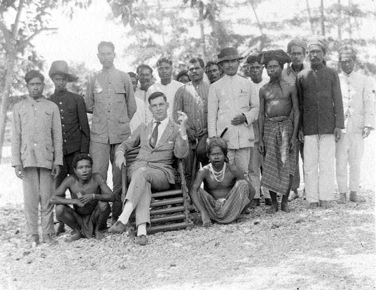 A white man in a suit sits surrounded by men of colour in an outdoor setting.