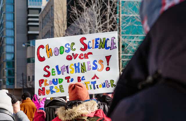 a woman in a crowd holds a sign reading CHOOSE SCIENCE OVER SELFISHNESS