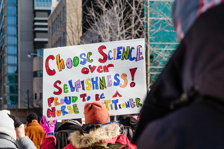 a woman in a crowd holds a sign reading CHOOSE SCIENCE OVER SELFISHNESS