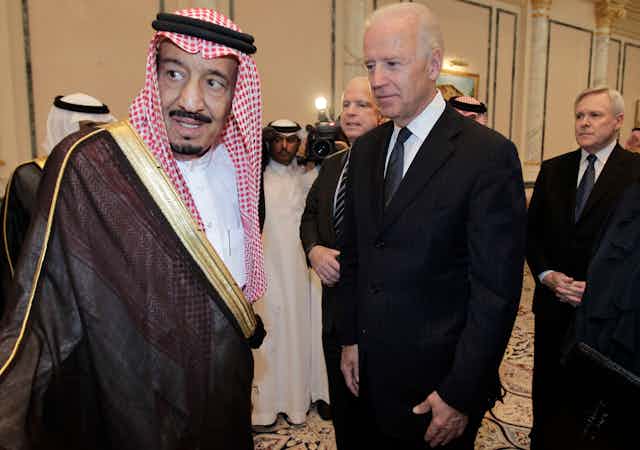 Joseph Biden in a black suit meets with King Salman bin Abdulaziz Al Saud in traditional Saudi dress.