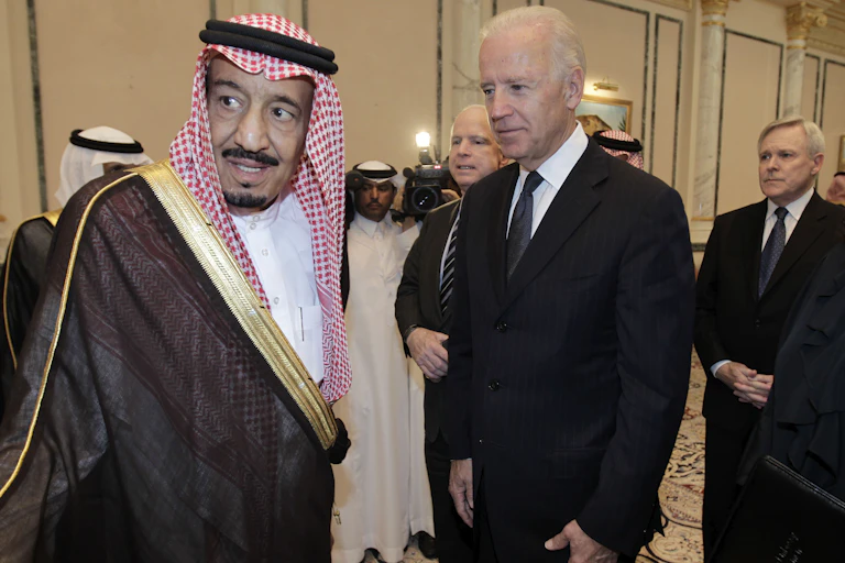 Joseph Biden in a black suit meets with King Salman bin Abdulaziz Al Saud in traditional Saudi dress.