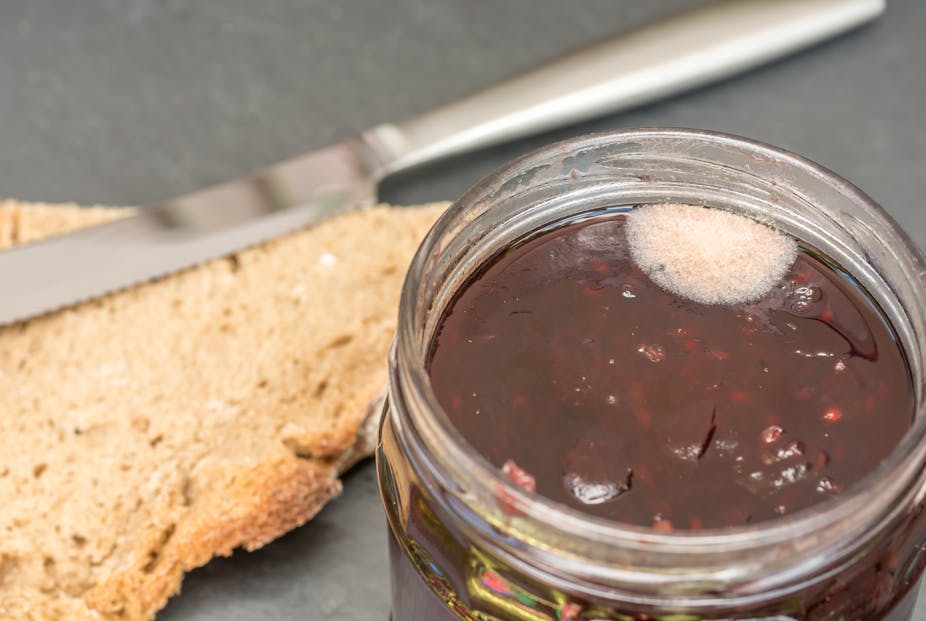 An open jar of jam with a spot of white mold sits next a crust of brown bread and a butter knife.