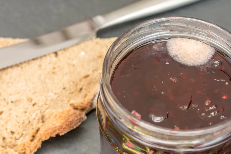 An open jar of jam with a spot of white mold sits next a crust of brown bread and a butter knife.