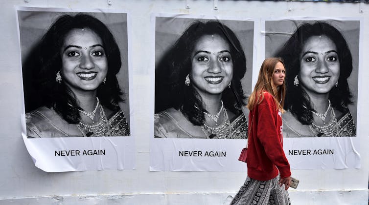 Three black and white photos show a young smiling woman who appears South Asian. Under her photo is a sign that says 'never again.' A  young woman walks past the street art.
