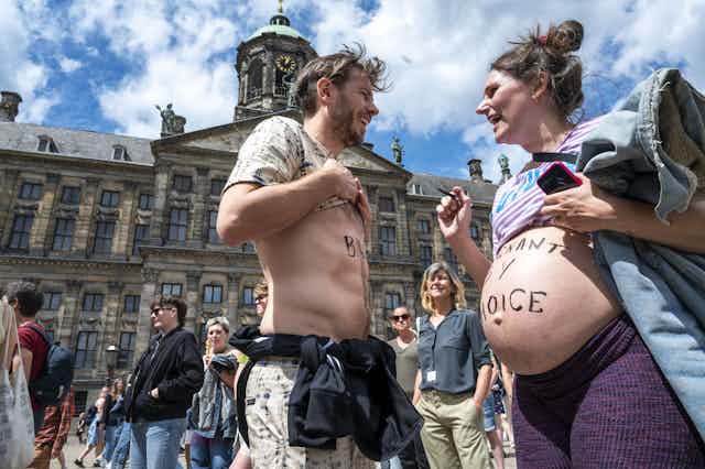 A man faces a pregnant woman, both have writing on their bodies about pro-choice - her stomach has writing that says, pregnant by choice. Behind them is an old looking European building. 