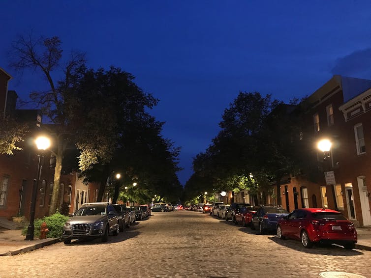Cars are parked on an old brick residential street at dusk with street lights and trees lining the sidewalks.