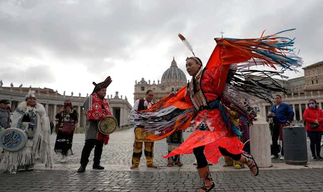 A dancer is seen in St. Peter's Square in regalia where the colour orange is dominant and a feather. 