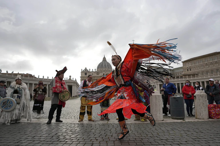 A dancer is seen in St. Peter's Square in regalia where the colour orange is dominant and a feather.