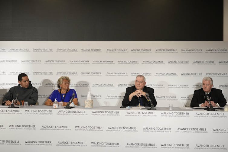 Four people are seen sitting in front of a backdrop that says 'walking together' and two bishops are wearing black clerical garments and collars.