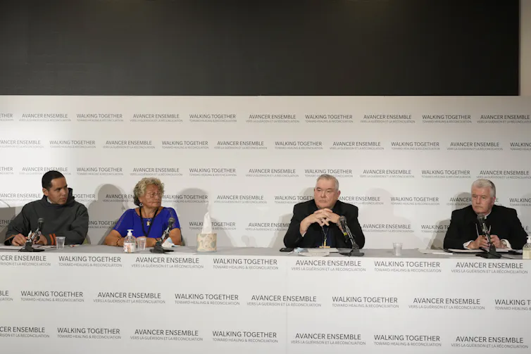 Four people are seen sitting in front of a backdrop that says 'walking together' and two bishops are wearing black clerical garments and collars.