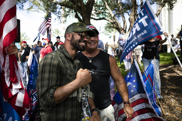 A brown-skinned man with a beard is pumping his fist as American flags and Trump banners wave in the background.