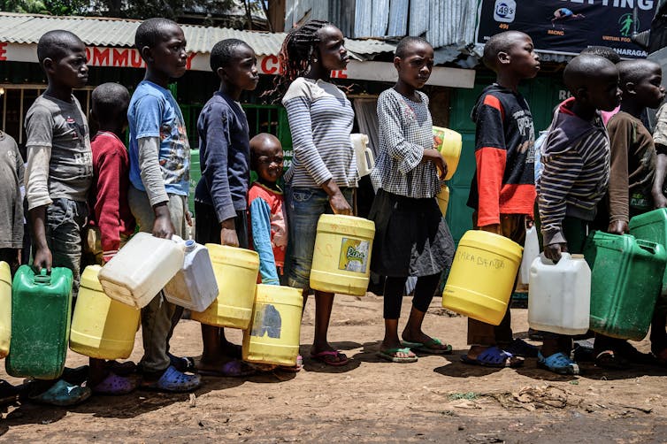 Children stand in line in a slum, carrying large plastic jugs.