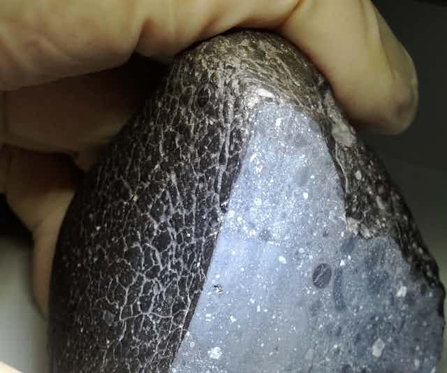 A photograph of a gloved hand holding a pyramid-shaped rock, blackish grey on the outside, sliced open to reveal white crystals inside.