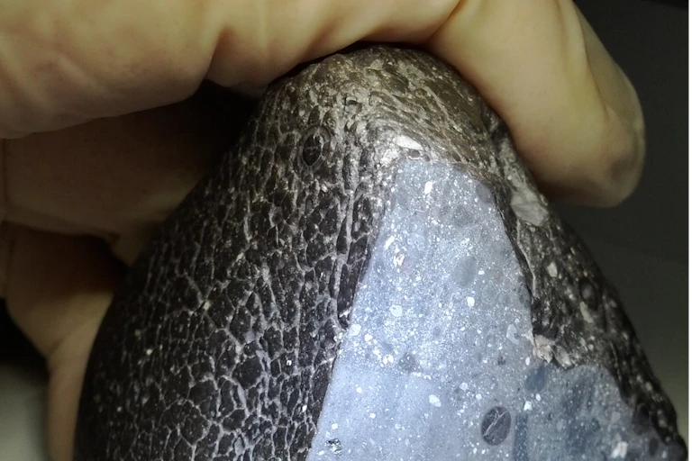 A photograph of a gloved hand holding a pyramid-shaped rock, blackish grey on the outside, sliced open to reveal white crystals inside.