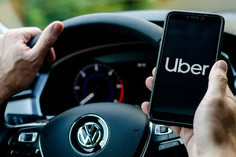 Driver's hands on steering wheel with one hand holding mobile phone with Uber on it