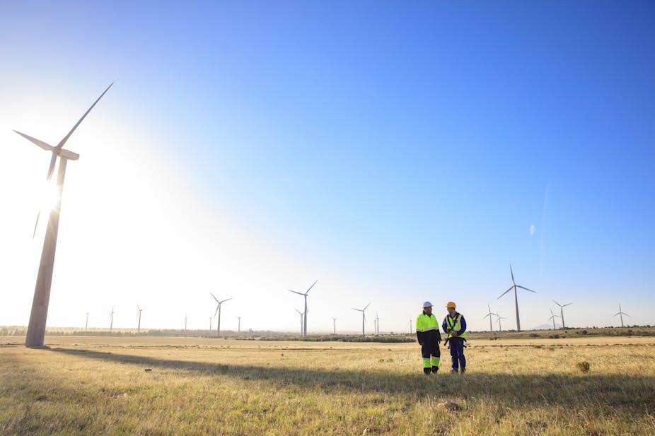Two engineers in work uniform on a wind farm with wind turbines in the background.