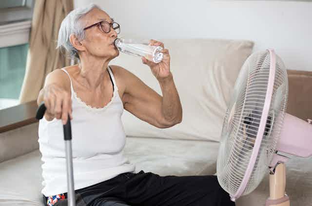 An older woman sits in front of a fan and drinks water.