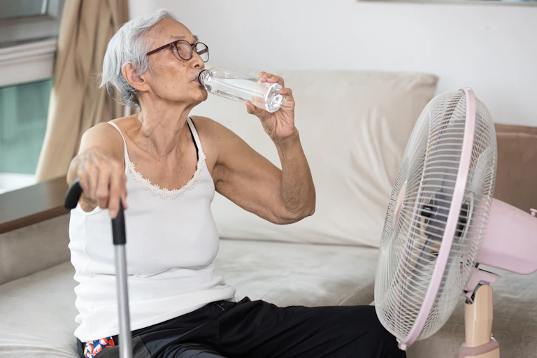 An older woman sits in front of a fan and drinks water.