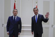 Two men in suits stand in front of flags in a white-panelled room.