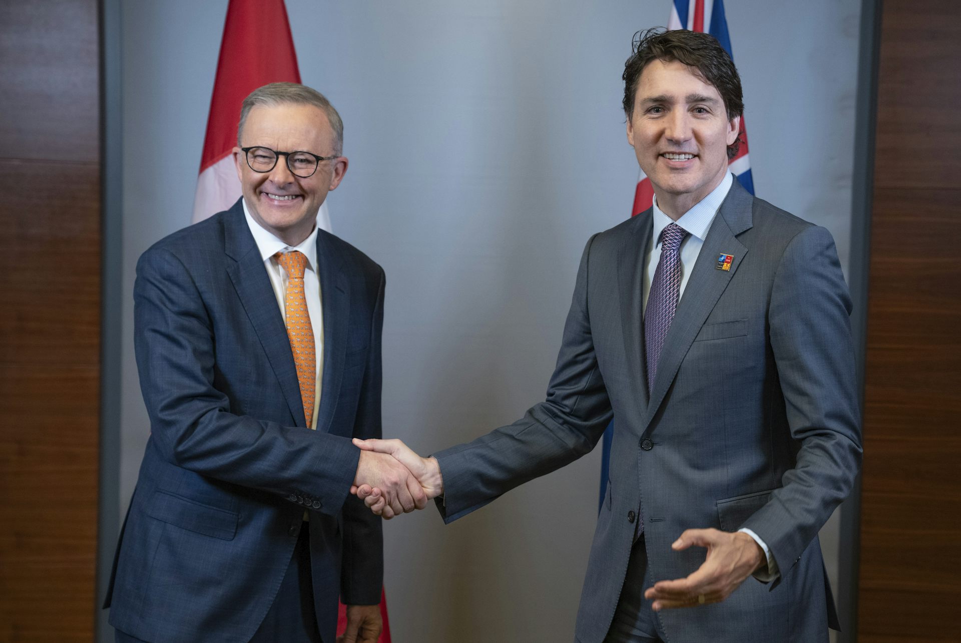 Two men in suits shake hands with flags behind them. Both are smiling.