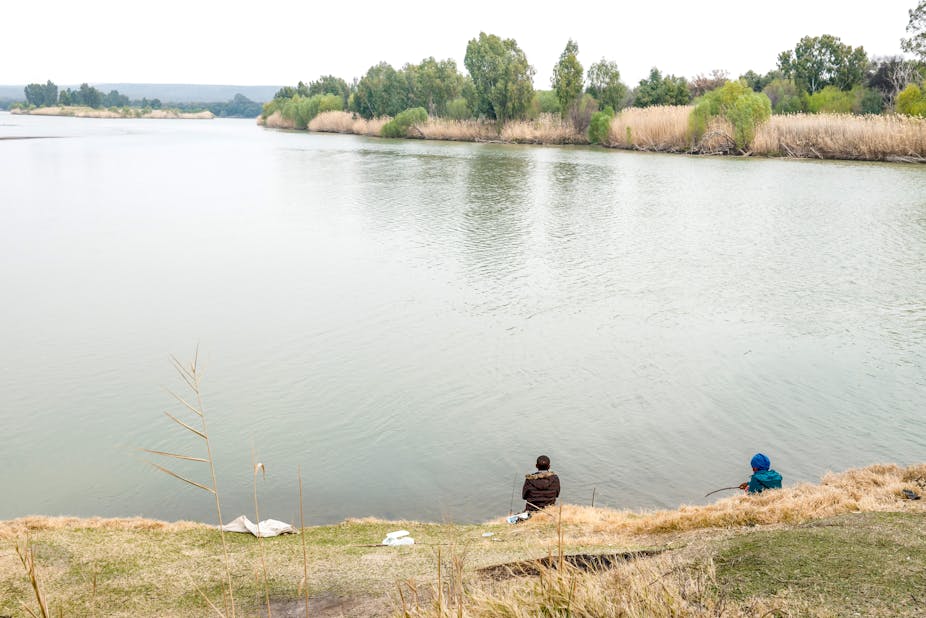 Two fishers, one in a dark jacket and the other in a blue jacket, sit with their lines cast into a still body of water. Trees stretch out in the distance and along the near banks of the river.