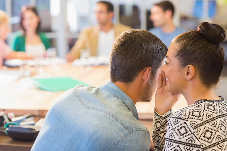 A woman whispers in her male colleague's ear as they are seated in a busy office