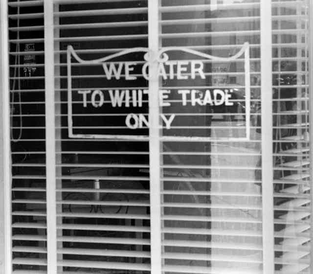 A sign with a curly frame in the window of a shop with the blinds open.