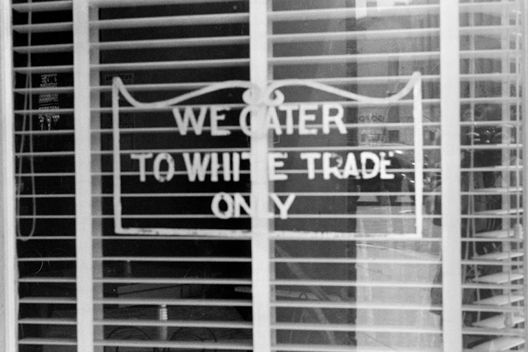 A sign with a curly frame in the window of a shop with the blinds open.
