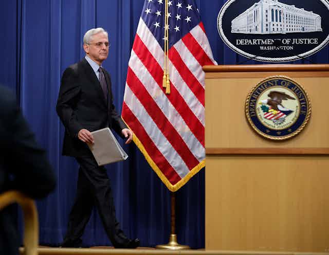 A man dressed in a business suit approaches a lectern bearing the seal of the U.S. Department of Justice near an American flag. 