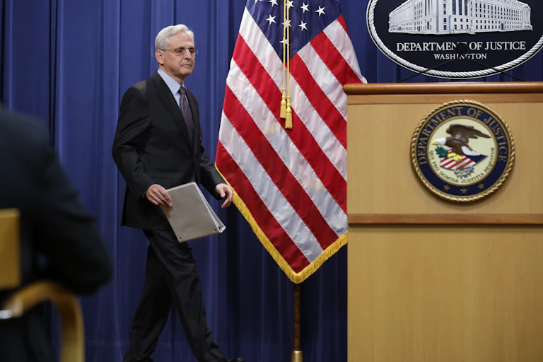 A man dressed in a business suit approaches a lectern bearing the seal of the U.S. Department of Justice near an American flag.