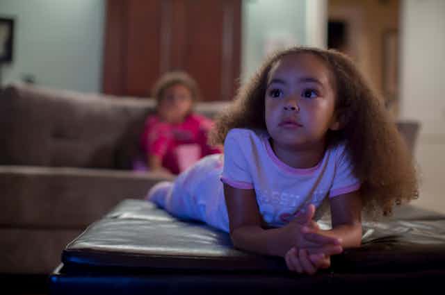 A young girl, resting on a sofa in her living room, watches TV.