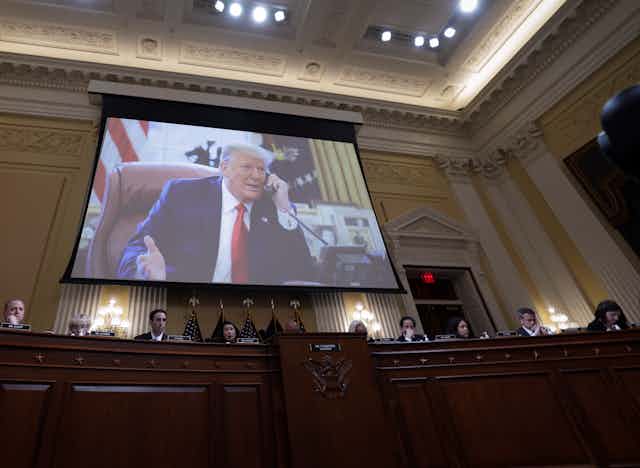 A white man dressed in a navy blue suit with a white shirt and red necktie appears on a video screen during a congressional hearing.