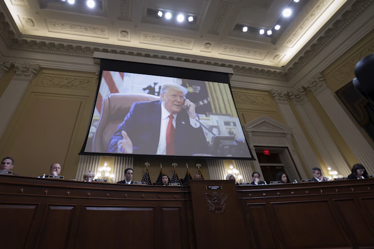 A white man dressed in a navy blue suit with a white shirt and red necktie appears on a video screen during a congressional hearing.
