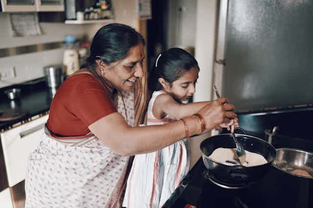 A grandmother standing behind her granddaughter, stirring a pot of food together.